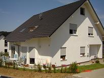 Biały, dwukondygnacyjny dom z ciemnym dachem, roletami w oknach i jasnymi elementami wykończeniowymi. Na pierwszym planie trawnik z młodymi roślinami, w tle widoczny fragment sąsiedniego budynku.